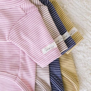 May include: Five striped baby onesies in pink, white, navy blue, yellow, and brown. The onesies have a small label that says "Sunny Days".