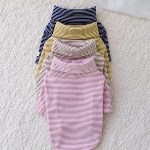 May include: Four striped turtleneck shirts for pets. The shirts are stacked on top of each other, with the navy blue shirt on top, followed by a yellow shirt, a beige shirt, and a pink shirt.
