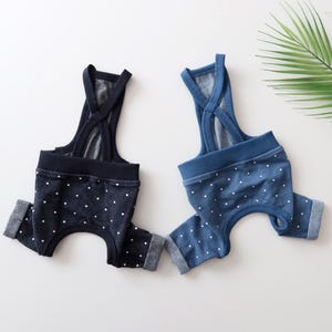 Puede incluir: Dos pares de petos para perros en denim azul marino y azul con lunares blancos. Los petos tienen un diseño de espalda cruzada y puños enrollados. El fondo es blanco con una hoja verde.