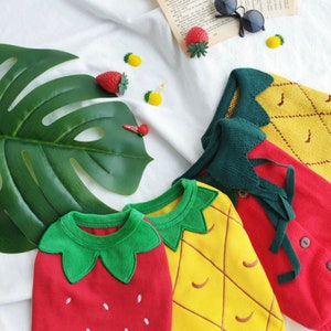 Pode incluir: Tr&ecirc;s conjuntos de malha para beb&eacute; com designs de frutas. Um morango vermelho, um anan&aacute;s amarelo e um anan&aacute;s amarelo com folhas verdes.