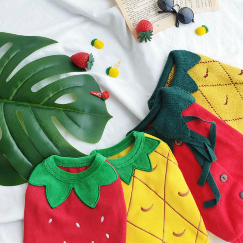 Pode incluir: Tr&ecirc;s conjuntos de malha para beb&eacute; com designs de frutas. Um morango vermelho, um anan&aacute;s amarelo e um anan&aacute;s amarelo com folhas verdes.