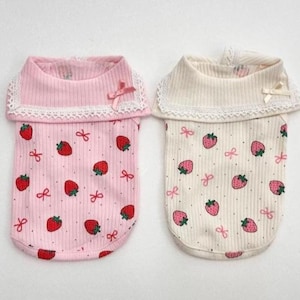 May include: Two dog shirts with a strawberry pattern. One is pale pink with red strawberries and bows, the other is off-white with pink strawberries and bows. Both have a ribbed collar with lace trim and a small bow.