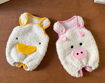 May include: Two plush pet costumes: one shaped like a duck with a yellow beak and trim, and the other like a pig with a pink snout and ears. Both are white and fluffy, designed for small pets.