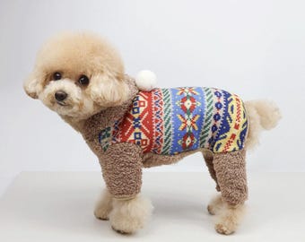 May include: A small brown and white dog wearing a brown and multi-colored patterned sweater with a white pom-pom on the hood.
