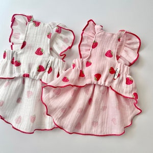May include: Two sleeveless dresses with ruffled straps and a strawberry print. One dress is white with red strawberries and the other is pink with red strawberries. Both dresses have a ruffled hem.