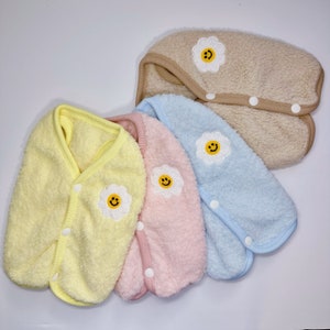 May include: Four plush dog vests in yellow, pink, blue, and beige with a smiling daisy embroidered on each. The vests have snap closures.