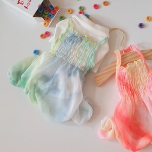 May include: Two pastel tie-dye dresses with ruffled straps. The dress on the left is light blue, green, and yellow. The dress on the right is pink, orange, and yellow.