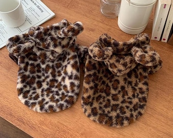 May include: Two leopard print dog sweaters with hoods. The sweaters are brown, tan, and black, with a soft, plush texture. The hoods have ear-like accents. The sweaters are designed to fit small dogs.