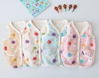 May include: Four sleeveless fleece vests in beige, white, light blue, and pink, each featuring a colorful cloud pattern, a front pocket, and snap button closures. Above the vests, a coloring book and five small gingerbread-shaped crayons.