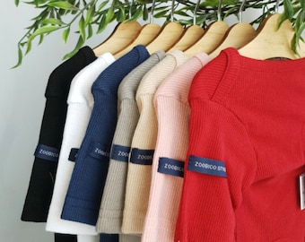 May include: A row of ten long-sleeved shirts hanging on wooden hangers. The shirts are in various colors, including black, white, navy blue, gray, beige, pink, and red. Each shirt has a small blue label with the text "ZOOBICO" on the sleeve.