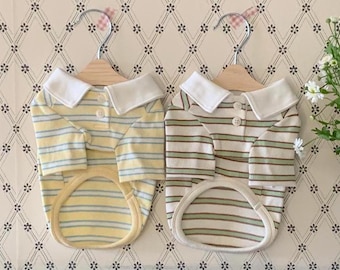 May include: Two dog polo shirts on wooden hangers. One is yellow with blue stripes, the other is white with green and brown stripes. Both have white collars and button closures. The shirts are displayed against a patterned background.