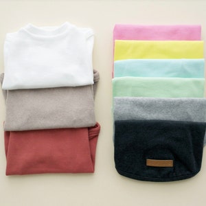 Pode incluir: Uma pilha de t-shirts dobradas em várias cores, incluindo branco, bege, vermelho e preto. As t-shirts estão dobradas cuidadosamente e dispostas em uma fila.