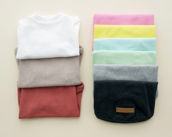 May include: A stack of folded t-shirts in various colors, including white, beige, red, and black. The shirts are folded neatly and arranged in a row.