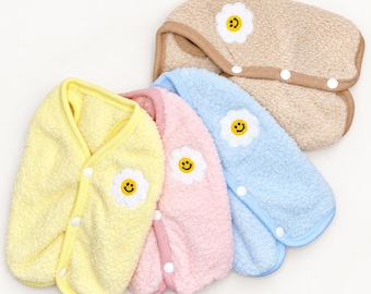 May include: Four sleeveless vests made of soft, fuzzy fleece material in yellow, pink, light blue, and beige. Each vest features a white embroidered flower patch with a yellow smiley face center and three white snap buttons along the front edge.