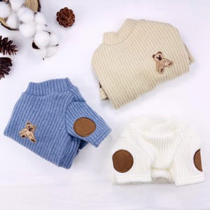 May include: Three pet sweaters in light blue, cream, and white. Each sweater has a ribbed texture, a small teddy bear patch, and brown elbow patches. The sweaters are folded and displayed on a white surface.