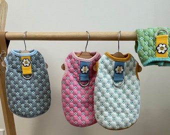 May include: Four quilted dog harnesses with a daisy design and a smiley face. The harnesses are in blue, pink, green, and white.