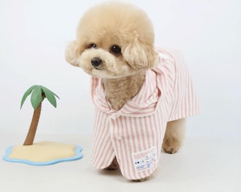 Puede incluir: Un perro pequeño, de color marrón claro, que lleva una camisa con capucha a rayas rosas y blancas. El perro está de pie junto a una pequeña isla decorativa con una palmera. La camisa tiene una pequeña etiqueta en la parte inferior.
