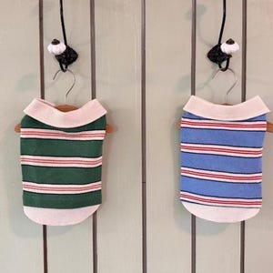 May include: Two sleeveless pet shirts hanging on small wooden hangers. One shirt is green with horizontal white and red stripes, and the other is blue with horizontal white and red stripes. Both shirts feature a solid white ribbed collar and hem.