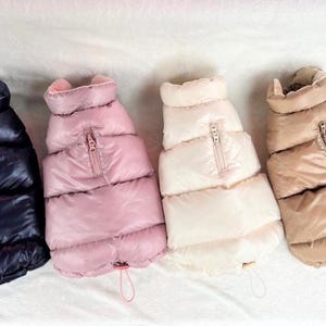 May include: Four quilted dog vests in navy, pink, off-white, and tan. Each vest features a zip closure and a drawstring hem. These vests are designed to keep dogs warm and comfortable.