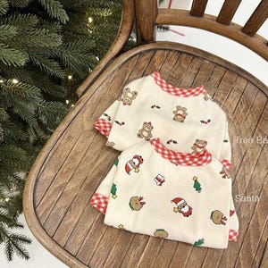 May include: Two cream-colored pajamas with red and white gingham trim. One pajama top features a print of bears and Christmas trees. The other pajama top features a print of Santa Claus, Christmas trees, and bears. The text "Tree Bear" and "Santa" is visible on the pajamas.