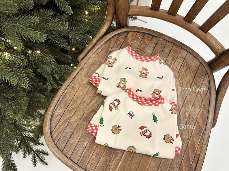 May include: Two cream-colored pajamas with red and white gingham trim. One pajama top features a print of bears and Christmas trees. The other pajama top features a print of Santa Claus, Christmas trees, and bears. The text "Tree Bear" and "Santa" is visible on the pajamas.
