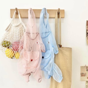 May include: A light pink and light blue mesh jumpsuit with a hood and pockets hangs on a wooden rack. A white mesh bag filled with lemons and a green fruit hangs to the left. A beige tote bag hangs to the right.