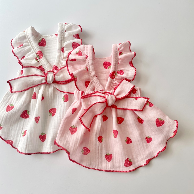 May include: Two sleeveless dresses with a strawberry print. One dress is white with a red bow and the other is pink with a red bow.