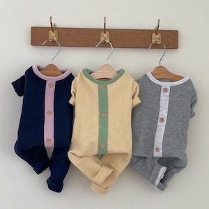 May include: Three baby onesies hanging on hangers. The onesies are made of a soft, ribbed fabric and have a contrasting color panel down the front. The onesies are in navy blue with a pink panel, yellow with a green panel, and gray with a white panel.