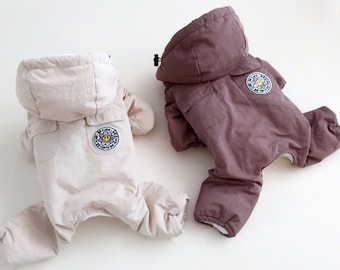May include: Two dog coats, one beige and one brown, with a smiling flower patch on the back. Both coats have hoods and are made of a soft, quilted fabric.