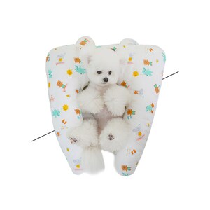 May include: A white dog with a fluffy tail is sitting in a white and green patterned dog bed. The bed is shaped like a U and has a raised edge. The dog is looking at the camera.