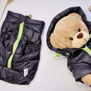 Puede incluir: Chaqueta acolchada negra con cremallera verde lima y un osito de peluche que lleva la chaqueta. La chaqueta tiene capucha y un pequeño logotipo en la parte inferior.