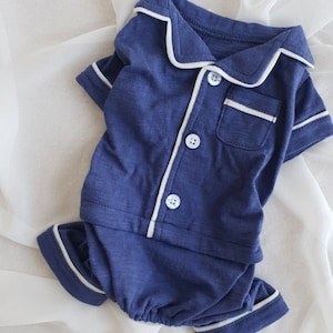 May include: A navy blue pajama set with white piping. The top has a button-up front and a pocket on the chest. The bottom is a pair of shorts.