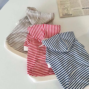 May include: Three striped hooded shirts for pets. The shirts are in grey, red, and blue with white stripes. The shirts have a pocket on the front.