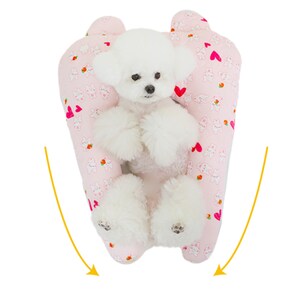 May include: A white dog sitting in a pink and white patterned dog bed with hearts and bunnies. The bed is shaped like a U and the dog is sitting in the middle.