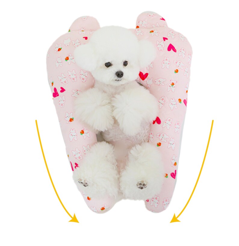 May include: A white dog sitting in a pink and white patterned dog bed with hearts and bunnies. The bed is shaped like a U and the dog is sitting in the middle.