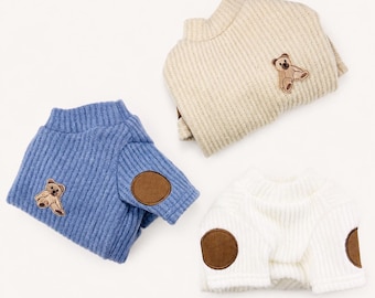 May include: Three ribbed sweaters in blue, beige, and white. Each sweater features a small embroidered teddy bear patch on the chest. The blue sweater includes brown circular elbow patches on the sleeves, while the white sweater also has brown patches.