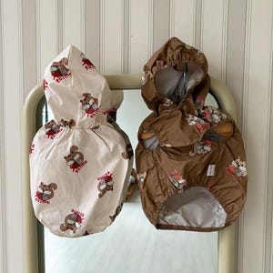 May include: Two hooded pet raincoats. One is cream-colored with a repeating pattern of squirrels and the word "taime" in red. The other is brown with the same squirrel pattern. Both have elastic waistbands.