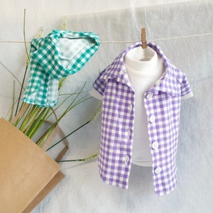 May include: Two checkered shirts, one green and one purple, hanging on a clothesline. The shirts are both short-sleeved and have a button-up front.