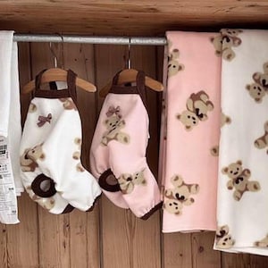 May include: White and pink baby outfits with teddy bear designs hang on wooden hangers. Two blankets with teddy bear patterns are also displayed. A white towel with red stripes hangs on the left.