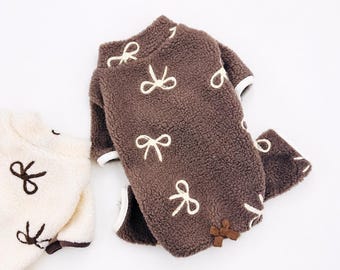 May include: A brown fleece pet sweater featuring a repeating pattern of white embroidered bows, a high collar, and a small brown ribbon bow detail at the hem. A second cream-colored sweater with brown bows is partially visible in the corner.