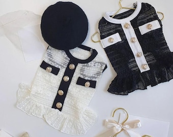 May include: Two dog dresses and a beret. One dress is white with black trim and gold buttons, the other is black with white trim and gold buttons. A black beret sits next to the white dress. Both dresses are on gold hangers.