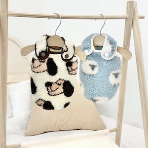 Pode incluir: Dois coletes de lã para animais de estimação com tema de cão e ovelha pendurados em um rack de madeira. O colete à esquerda é branco com rostos de cães marrons e o colete à direita é azul claro com rostos de ovelhas brancas.