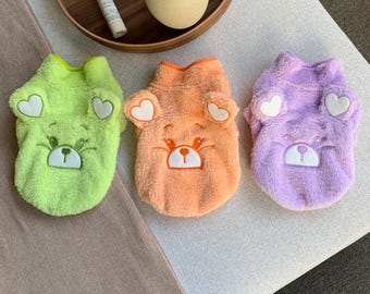 May include: Three plush dog sweaters in green, orange, and purple with embroidered bear faces and hearts on the ears.