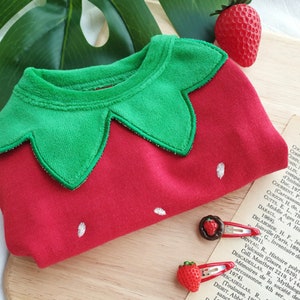 Pode incluir: Macac&atilde;o de beb&eacute; vermelho e verde com um design de morango. O macac&atilde;o tem um colar de feltro verde em forma de folha de morango e um corpo de feltro vermelho com uma semente de morango bordada em branco.