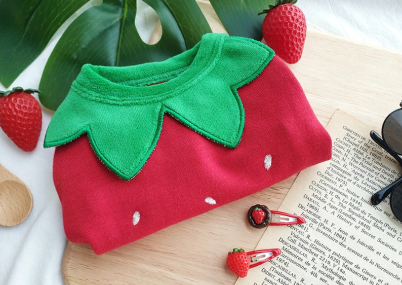 Pode incluir: Macac&atilde;o de beb&eacute; vermelho e verde com um design de morango. O macac&atilde;o tem um colar de feltro verde em forma de folha de morango e um corpo de feltro vermelho com uma semente de morango bordada em branco.