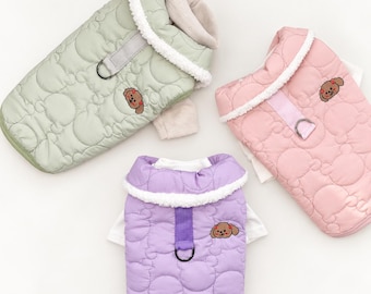 May include: Three quilted pet vests in light green, soft pink, and lavender. Each vest features a white sherpa-lined collar, a D-ring attachment on a vertical strap, and an embroidered brown puppy face patch on the quilted fabric surface.