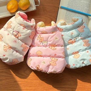 May include: Three dog puffer vests in white, pink, and light blue, each adorned with a repeating cartoon bear pattern. The vests have a quilted design, a metal D-ring, and a contrasting collar. The vests are designed for small dogs.