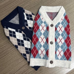May include: Two argyle pattern sweaters. One is navy blue with tan and white diamonds. The other is a cream-coloured cardigan with red and light blue diamonds. Both sweaters have a V-neck and button closures.
