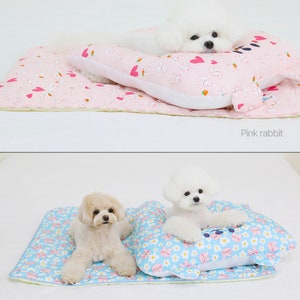 May include: Two white dogs are laying on a bed with a pillow. The bed is pink with a white rabbit pattern and the pillow is pink with a white rabbit pattern. The other bed is blue with a white rabbit pattern and the pillow is blue with a white rabbit pattern. The text "Pink rabbit" is below the first bed and the text "Blue rabbit" is below the second bed.