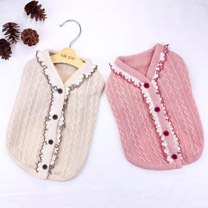 May include: Two dog sweaters, one cream-colored and one pink, are displayed. Both have a cable knit design, ruffled trim, and button closures. The cream sweater is on a wooden hanger with "NK pup" printed on it.